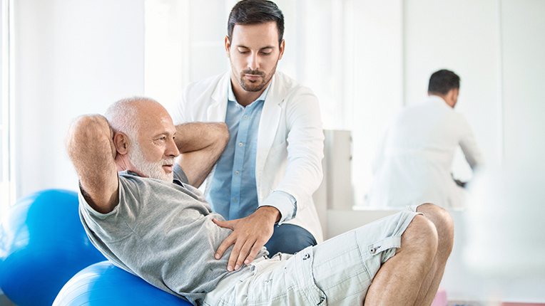 Closeup side view of an early 60's senior gentleman having physical therapy appointment. Mid 30's expert doctor is helping the man do some light exercise. He's doing some crunches while sitting on a blue fitness ball.